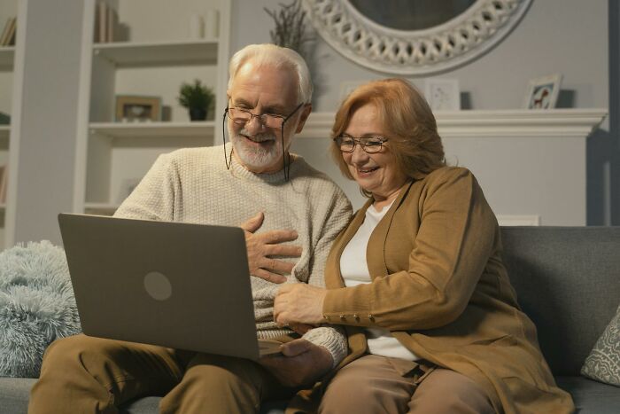 Elderly couple smiling and sharing uplifting stories of progress and hope in America while using a laptop at home.