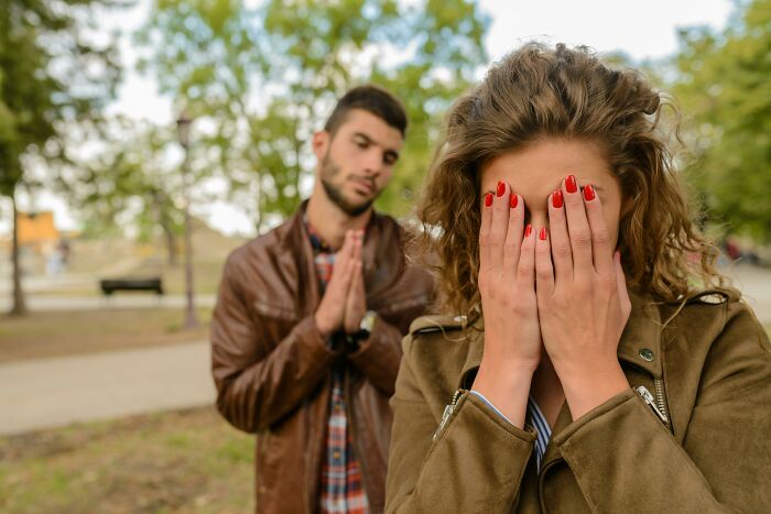 A woman covering her face with hands painted red nails while a man in a leather jacket gestures apologetically outdoors, reflecting relationships.