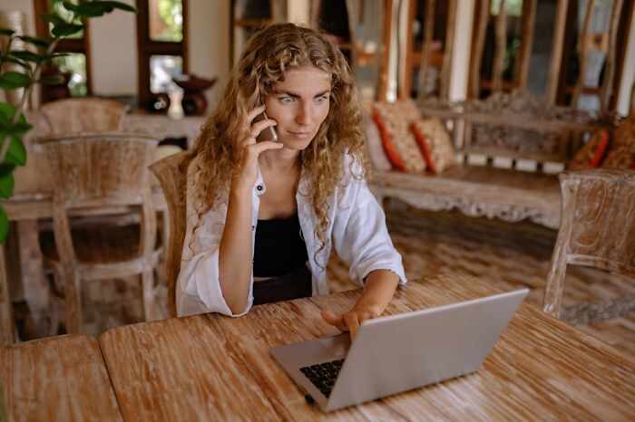 Young woman at wooden table looking concerned while texting on phone and using laptop, illustrating accidental texts causing panic.