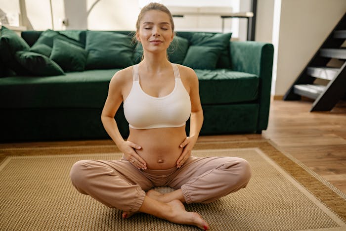 Pregnant woman meditating in living room, seated cross-legged on rug with hands on belly in a calm moment.