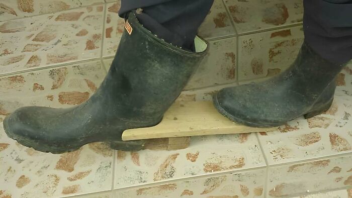 Person wearing black boots using a wooden stick to keep the boots elevated on tiled floor, Eastern European humor.