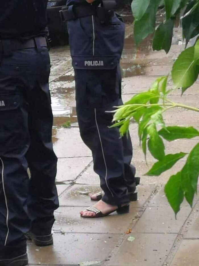 Eastern European police officers standing outside with one wearing open-toe sandals in uniform pants on a rainy pavement.
