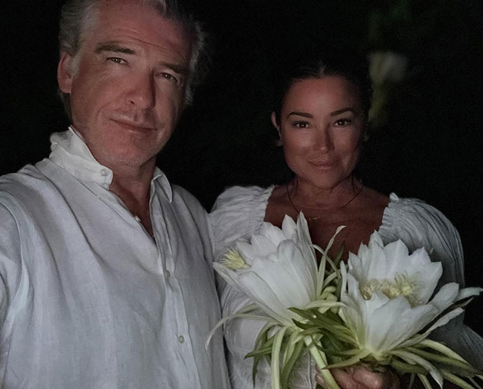 Pierce Brosnan and his wife in white outfits, with her holding large white flowers, amid online trolls discussing transformation.