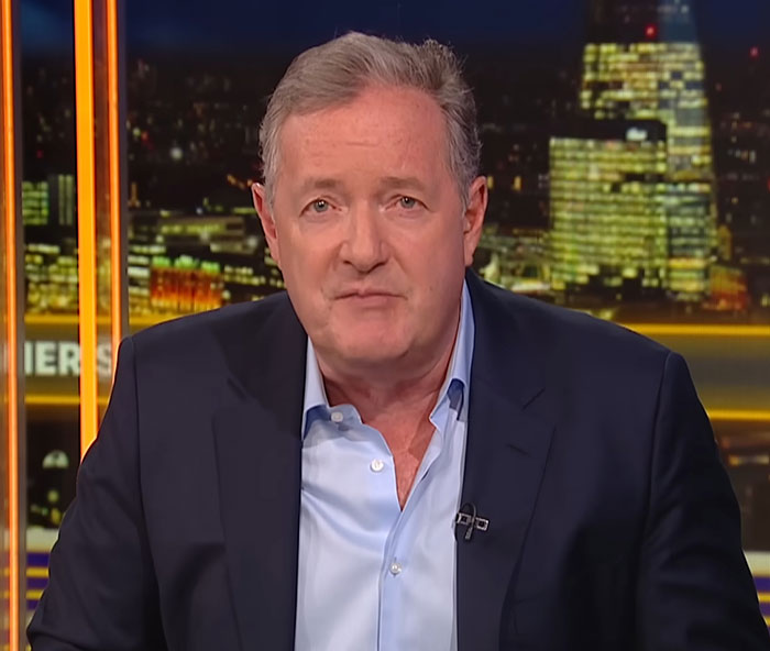 Piers Morgan wearing a suit and blue shirt, speaking on a nighttime news set with city lights in the background Piers Morgan wearing a suit and blue shirt, speaking on a nighttime news set with city lights in the background