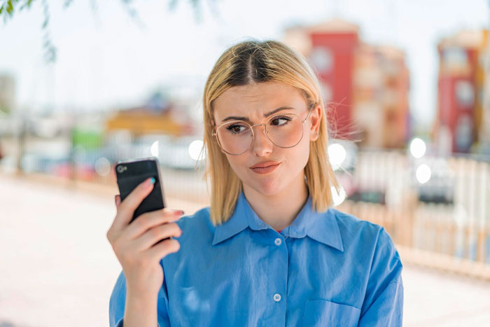 Young woman in glasses looking skeptical at her smartphone while standing outdoors on a sunny day. Young woman in glasses looking skeptical at her smartphone while standing outdoors on a sunny day.