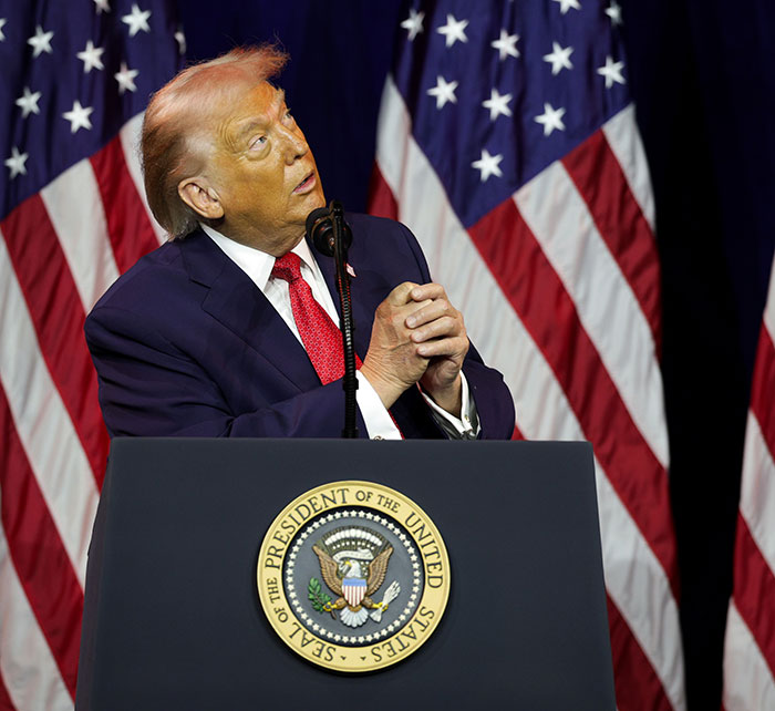 Former President Trump at a podium with multiple American flags, showing hair with a pinkish tint, styled professionally. Former President Trump at a podium with multiple American flags, showing hair with a pinkish tint, styled professionally.