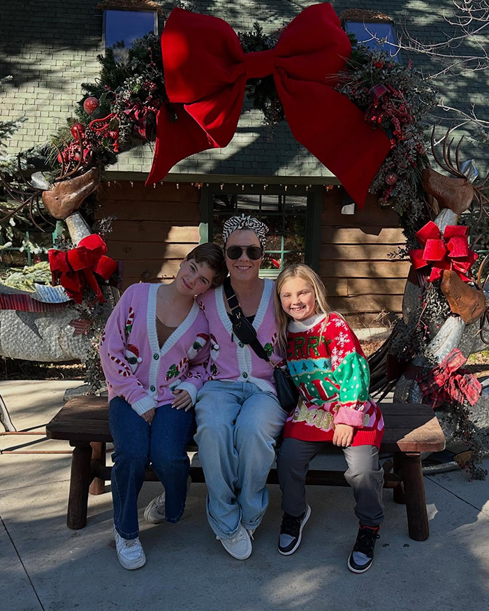 Pink with family sitting on a bench under a large festive wreath decorated with red bows and holiday ornaments. Pink with family sitting on a bench under a large festive wreath decorated with red bows and holiday ornaments.