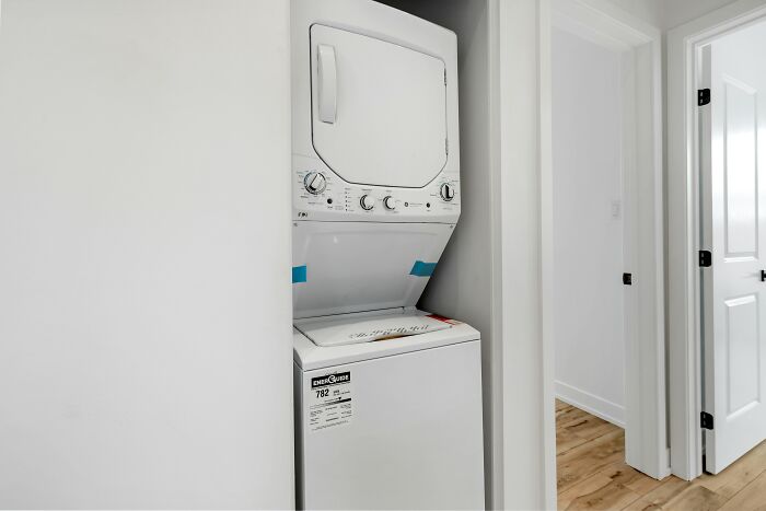 Stacked washer and dryer in a narrow laundry closet inside a modern home hallway with white walls and wooden floors.