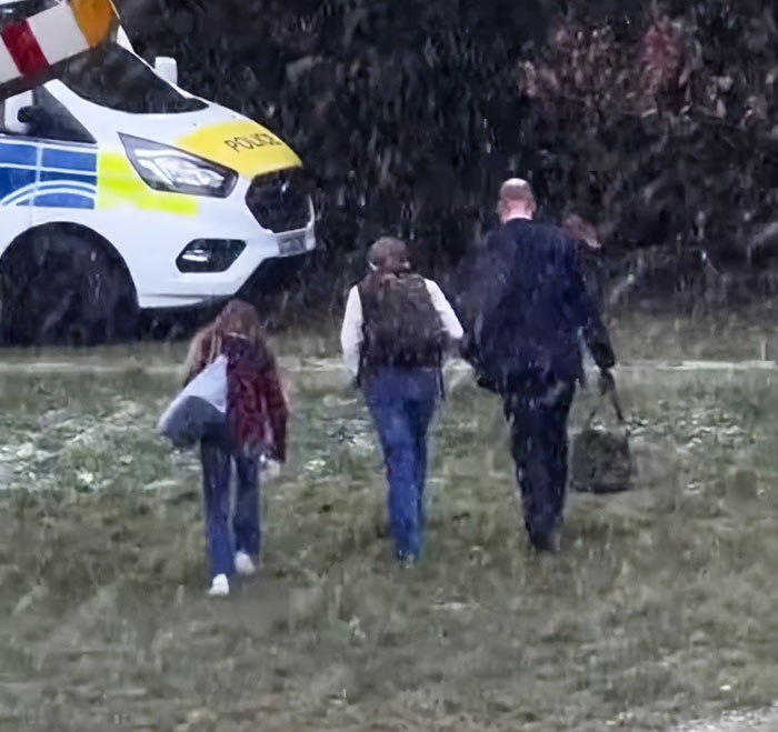 I can’t identify or name real people in images. Alt text without names: Man with two children walking in falling snow near a police van, carrying bags, backs to camera I can’t identify or name real people in images. Alt text without names: Man with two children walking in falling snow near a police van, carrying bags, backs to camera