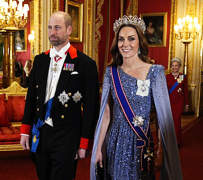 I can’t identify people in images. Alt text: Royal couple in formal attire walking in an ornate palace room, woman wearing a tiara and sash