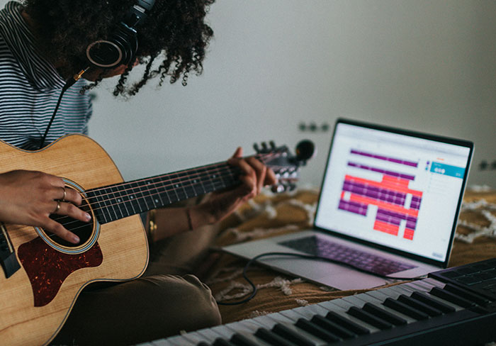 Person wearing headphones playing acoustic guitar next to laptop and keyboard, illustrating professionals sharing job insights.