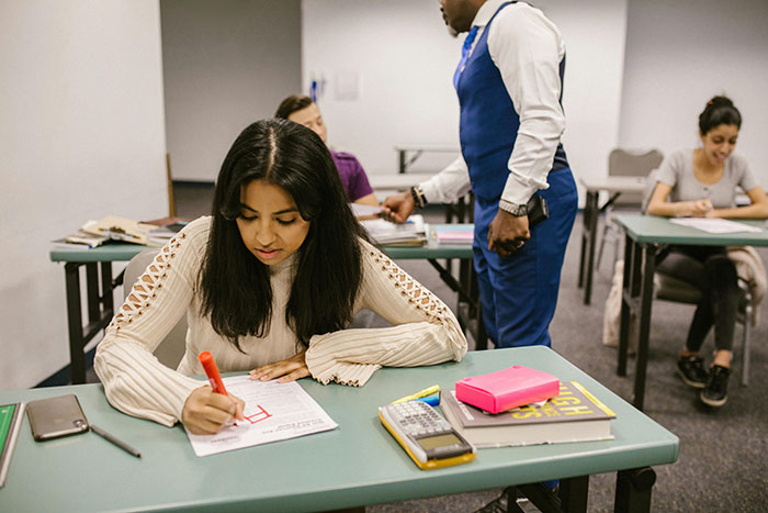 Student writing a test while professor enforces phone rule, highlighting students push back and risk of failing class.