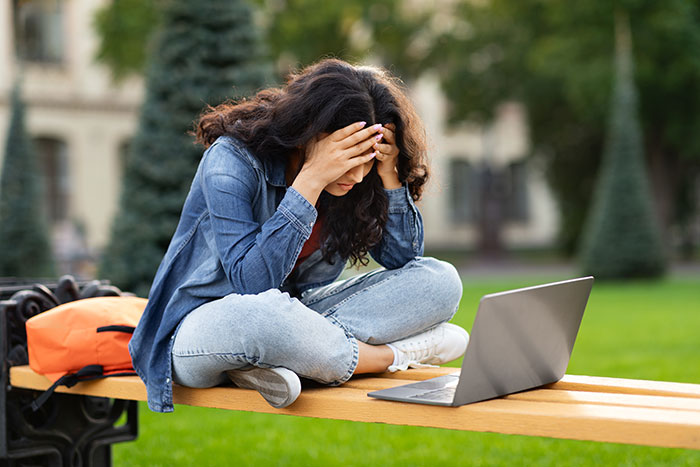 Stressed student sitting on bench outdoors with laptop, struggling after professor enforces phone rule without warning.