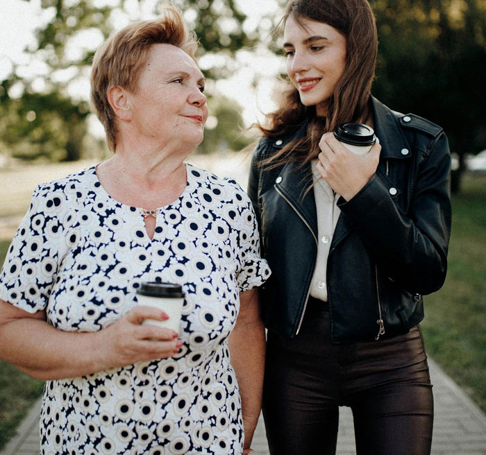 Mother-in-law and daughter-in-law walking outdoors, each holding a coffee cup, sharing a moment together.
