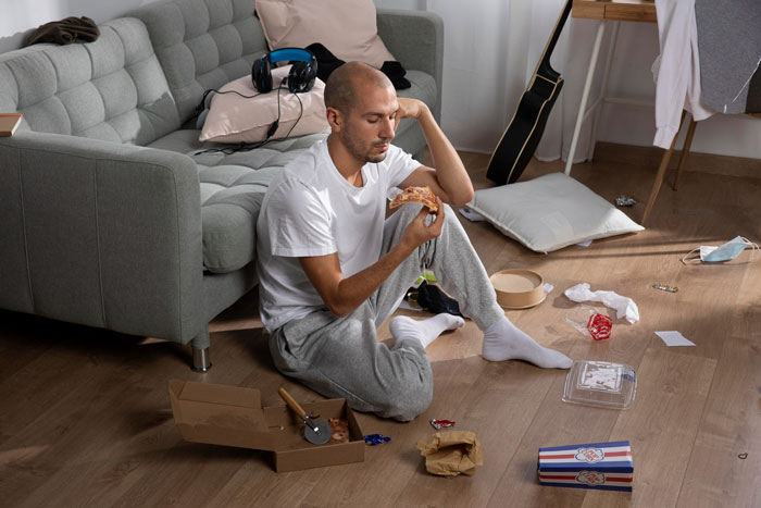 Man-child hubby sitting on messy floor eating pizza, surrounded by clutter in living room, reflecting working wife challenges.