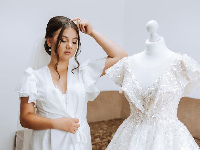 Young woman in a white robe with a veil preparing near a sparkling wedding dress on a mannequin indoors.
