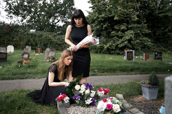 Two women in black mourning at a grave with flowers, reflecting loss and conflict over a daughter's wedding dress.