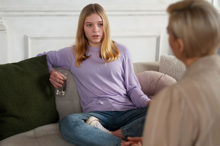 Teenage daughter sitting on couch holding a glass, talking to an older woman about a wedding dress dispute with her mom.