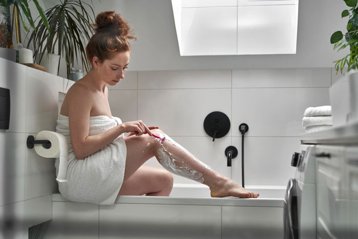 Young woman wrapped in a towel shaving her legs in a modern bathroom, addressing personal hygiene and grooming. Young woman wrapped in a towel shaving her legs in a modern bathroom, addressing personal hygiene and grooming.