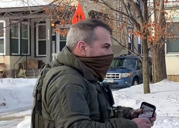 Man in tactical gear and brown face covering holding a phone outdoors near a snowy residential area during daylight.