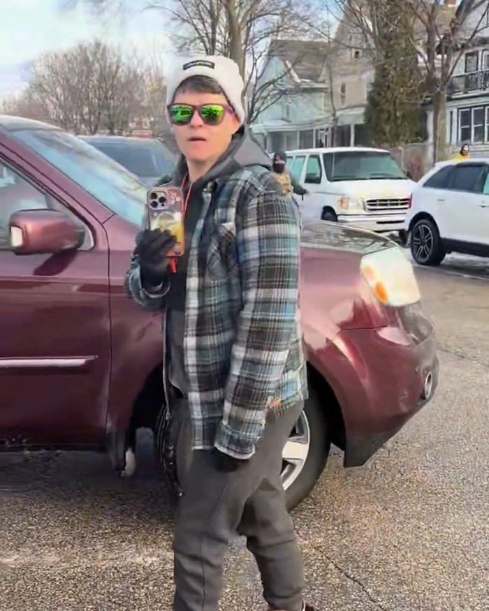 Person wearing green sunglasses and a hat, holding a smartphone near parked vehicles in an urban neighborhood scene. Person wearing green sunglasses and a hat, holding a smartphone near parked vehicles in an urban neighborhood scene.