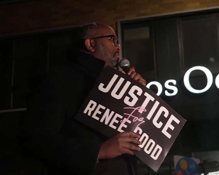 Man holding a Justice for Renée Good sign speaking into a microphone during a public gathering about fatal shooting context. Man holding a Justice for Renée Good sign speaking into a microphone during a public gathering about fatal shooting context.