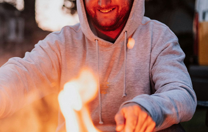 Man in gray hoodie smiling by a fire, capturing the moment of people getting the last laugh with glorious revenge.