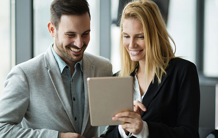 Two colleagues smiling and sharing a moment of glorious revenge on a tablet in a bright modern office.