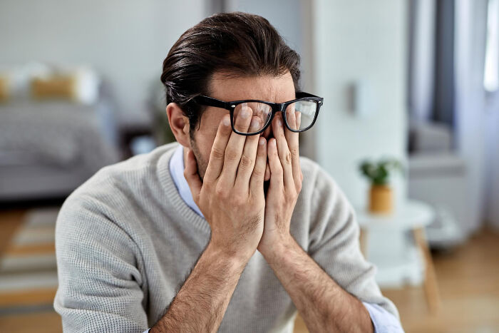 Man wearing glasses and a gray sweater covering his face indoors, reflecting on rich people norms and lifestyle insights.