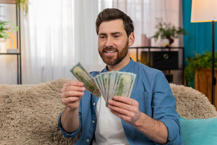 Man sitting on a couch smiling and holding cash, illustrating rich people norms shared by insiders on the 1% lifestyle.