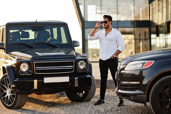 Man in sunglasses standing between luxury cars, illustrating rich people norms from inside info on the 1 percent.