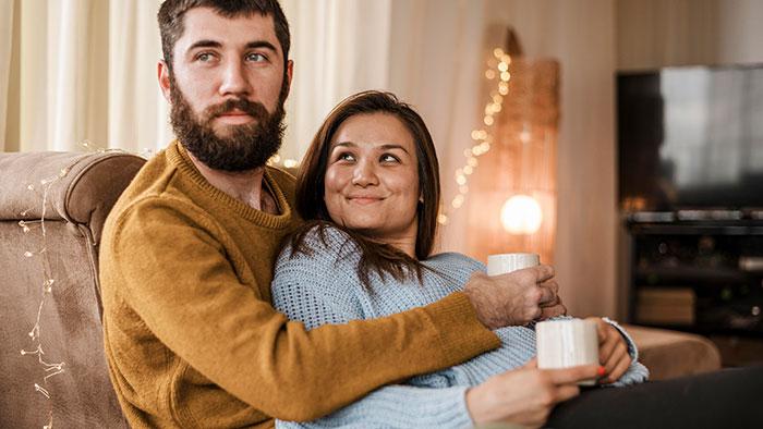 Couple sitting on couch holding mugs, woman looking at man with a smile in a cozy living room setting.
