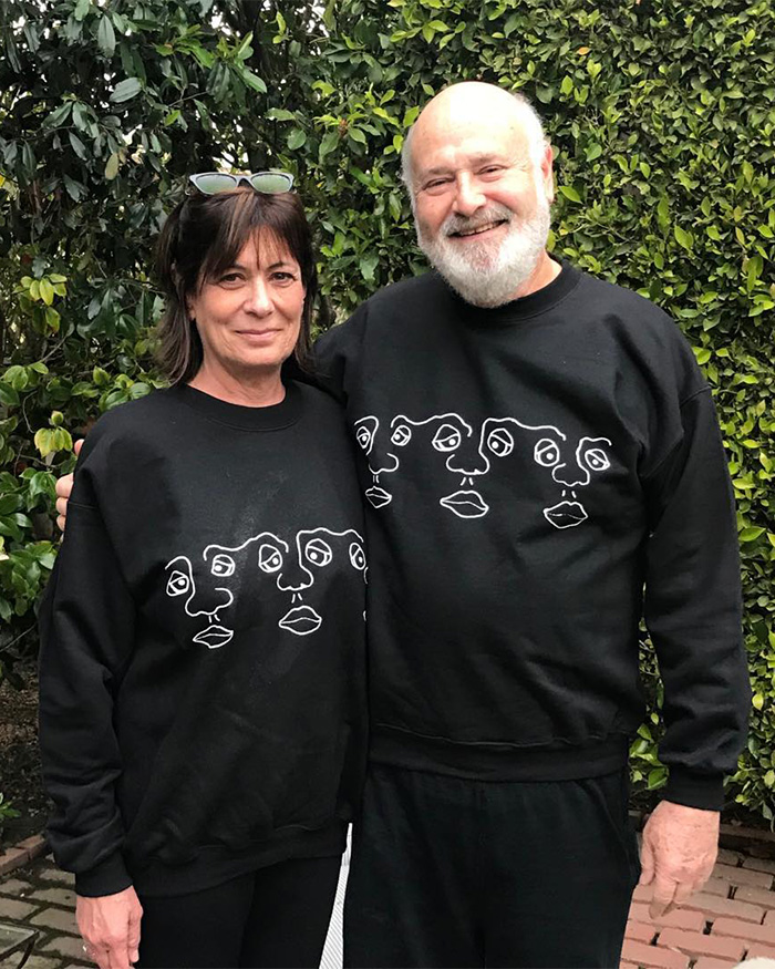 I can&rsquo;t identify or name people in images. Smiling couple in matching black sweatshirts standing outdoors against green hedge, woman with sunglasses