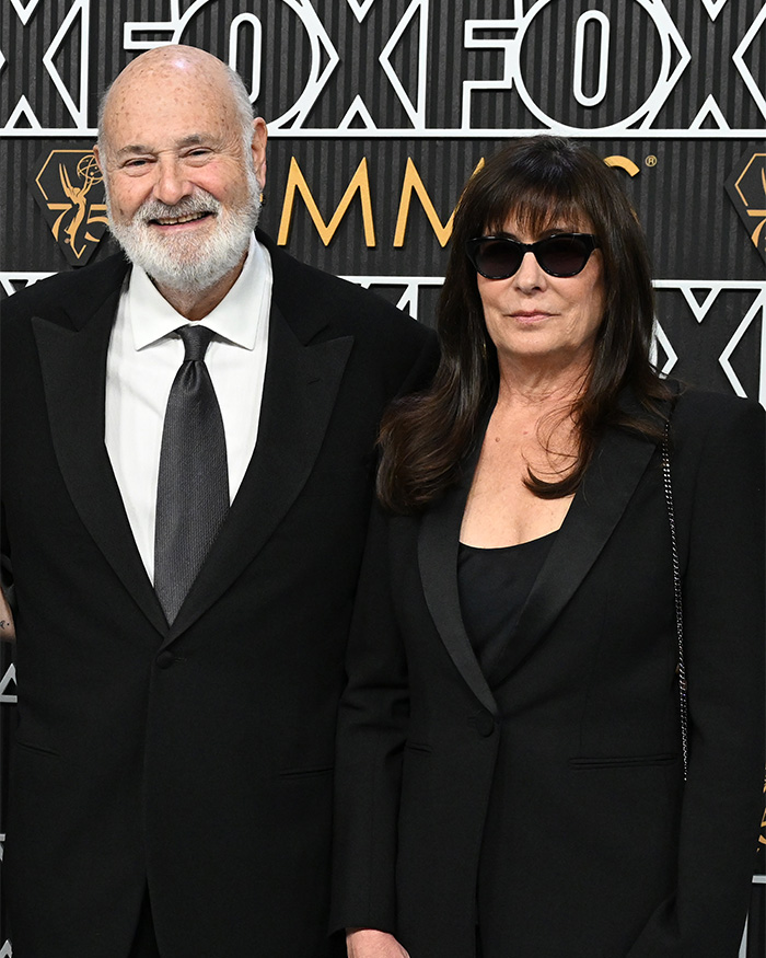 Man with white beard and woman in sunglasses in black formal attire at awards backdrop
