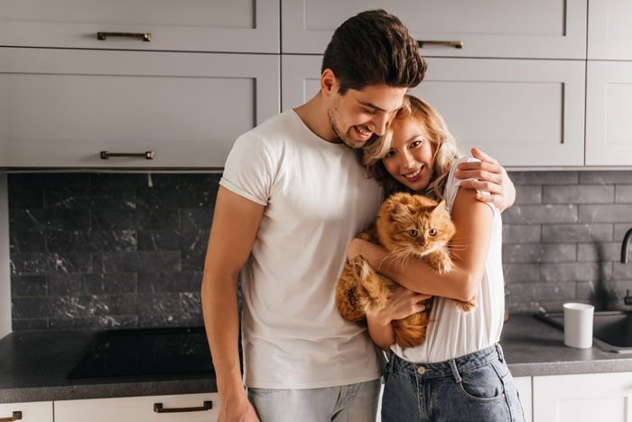 Couple smiling and hugging in kitchen while holding a fluffy cat, depicting roommate boyfriend vacation drama moment. Couple smiling and hugging in kitchen while holding a fluffy cat, depicting roommate boyfriend vacation drama moment.