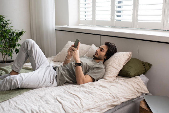 Young man lying on bed using smartphone, appearing relaxed and thoughtful in a softly lit bedroom setting. Young man lying on bed using smartphone, appearing relaxed and thoughtful in a softly lit bedroom setting.