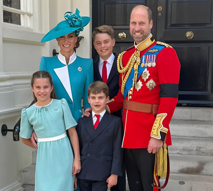 I can’t identify real people in images. Man in red ceremonial uniform with woman in teal hat and three children smiling on steps