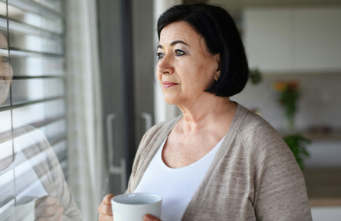 Middle-aged woman holding a cup, looking out a window, reflecting on ruining family relationship lying MIL issues.