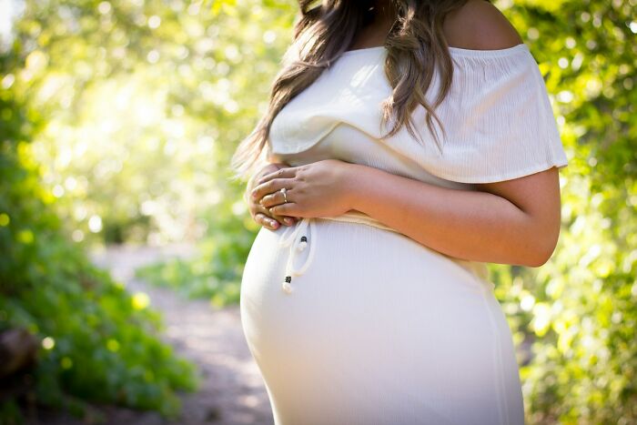 Pregnant woman in a white dress holding her belly outdoors in a natural setting with soft sunlight and greenery.
