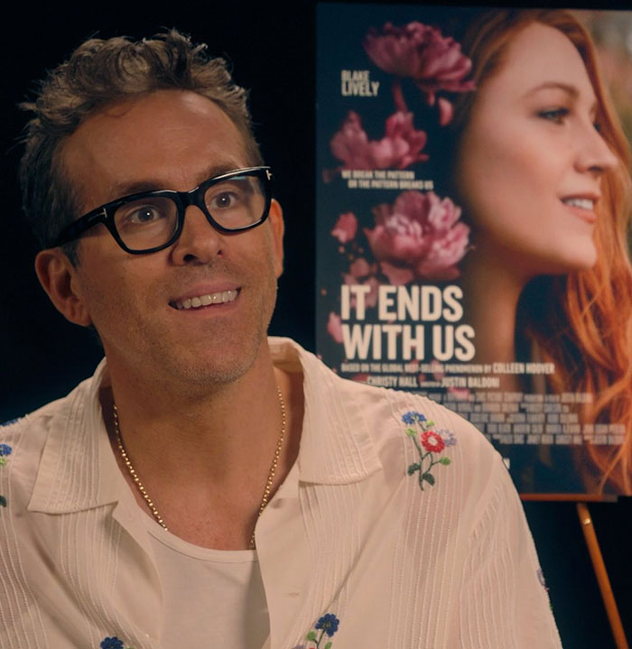 Ryan Reynolds wearing glasses and a white shirt during an interview with It Ends With Us movie poster behind him Ryan Reynolds wearing glasses and a white shirt during an interview with It Ends With Us movie poster behind him