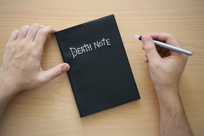 Hands holding a black notebook labeled Death Note on a wooden desk, hinting at high school controversies and unusual events.
