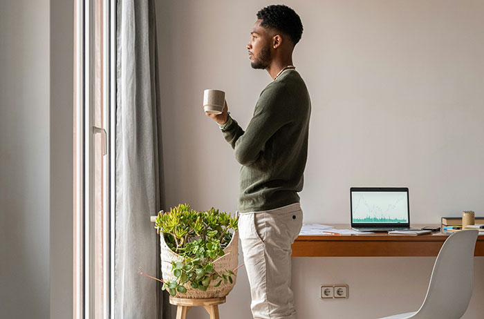 Man holding mug looking out a window by desk with laptop and plant, Saw Images On Google