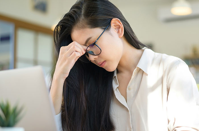 Woman rubbing temples at laptop, distressed after viewing images on Google, wearing glasses in bright workspace