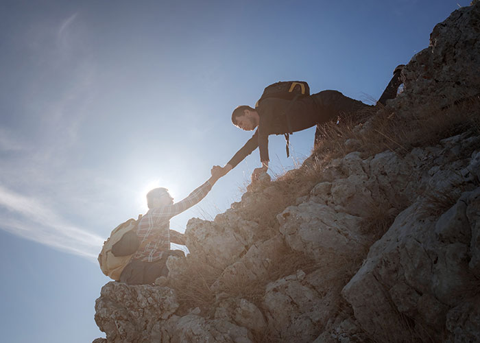 Two hikers experiencing a terrifying close call as one helps the other climb a steep rocky mountain trail.