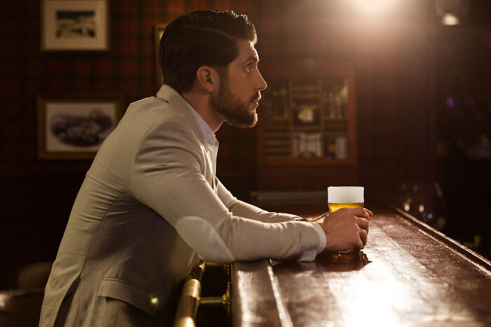 Man in a blazer sitting alone at a bar holding a beer, reflecting on meeting their high school bully as adults.