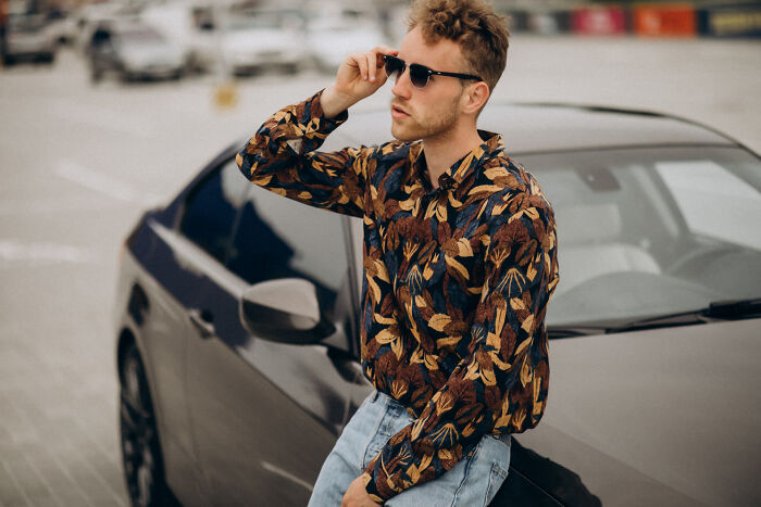 Young man wearing sunglasses and patterned shirt leaning on a car, symbolizing meeting high school bully as adults and karma.