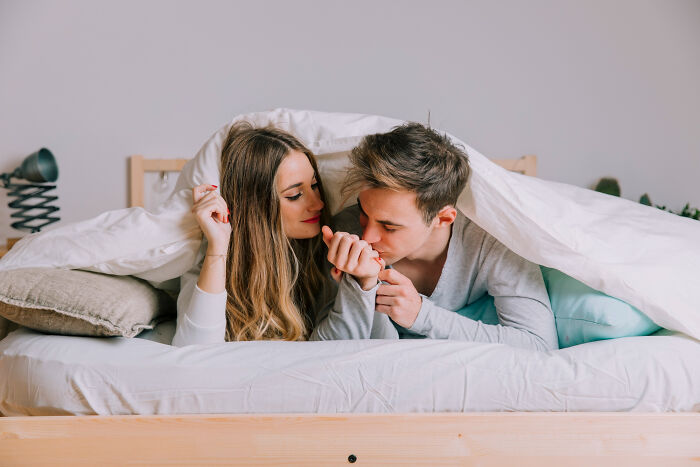 Young couple lying in bed under white covers, sharing a tender moment, illustrating adults meeting their high school bully karma.