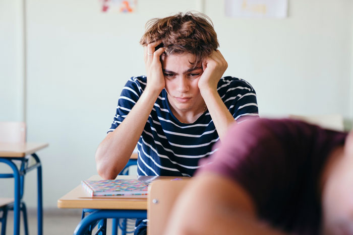 Stressed teenage boy sitting at desk in classroom, struggling with group shaming pushed by unhinged teacher. Stressed teenage boy sitting at desk in classroom, struggling with group shaming pushed by unhinged teacher.