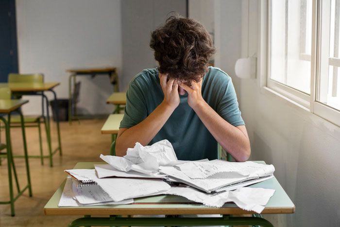 Teen boy in classroom stressed and overwhelmed with crumpled papers, highlighting impact of group shaming in school setting. Teen boy in classroom stressed and overwhelmed with crumpled papers, highlighting impact of group shaming in school setting.