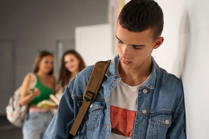 Teen boy in denim jacket looks down alone while two girls talk in background, illustrating group shaming in class scenario.